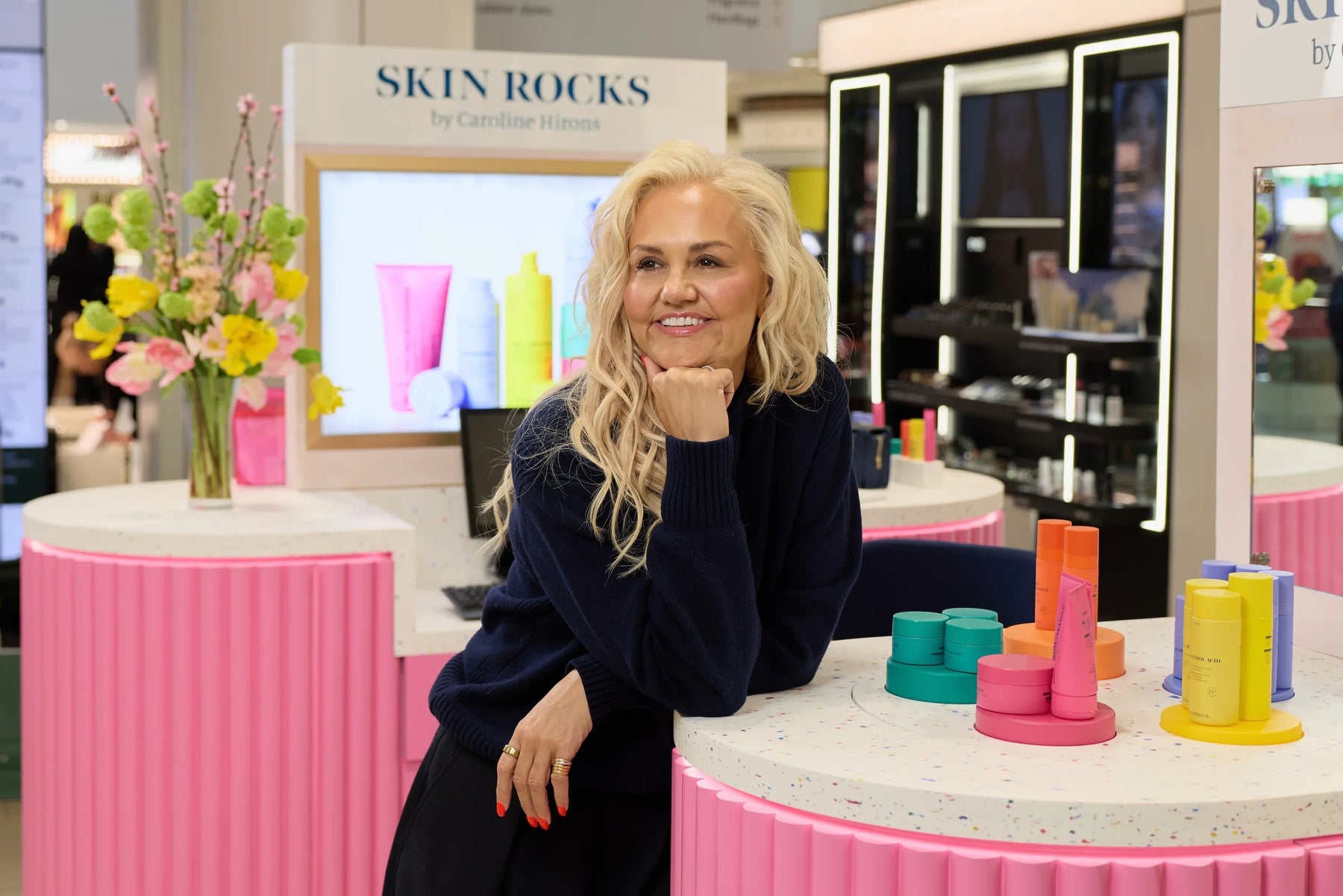 Skin Rocks by Caroline Hirons | Caroline Hirons smiles beside colorful skincare products at a bright pink retail display